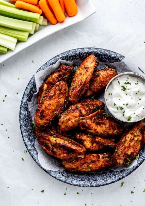 an overhead shot of crispy air fryer chicken wings in a bowl lined with parchment paper beside a dish of dipping sauce and celery and carrot sticks on a white plate in the corner of the shot.