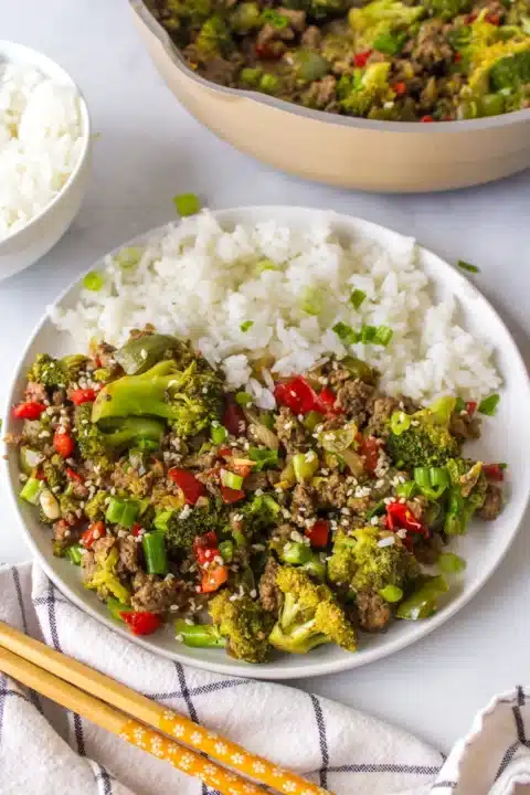 a white plate with white rice and a side of bison stir fry with broccoli on the side in the foreground you can see a napkin and a pair of chopsticks.