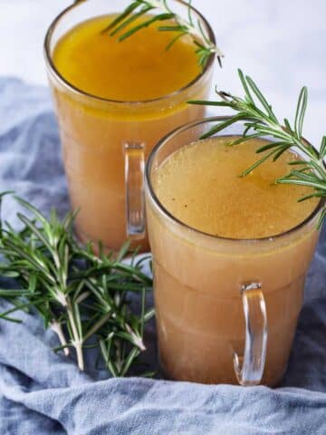 Two cups filled with broth and topped with fresh Rosemary leaves.