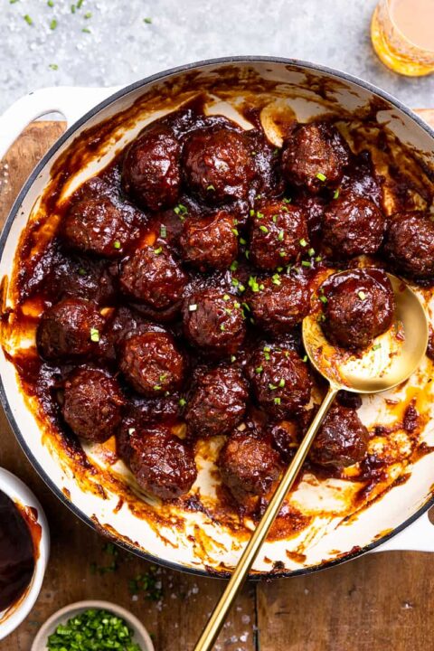 an overhead shot of bourbon bbq bison meatballs in a enameled cast iron skillet on a wooden surface with a golden spoon lifting up a meatball.