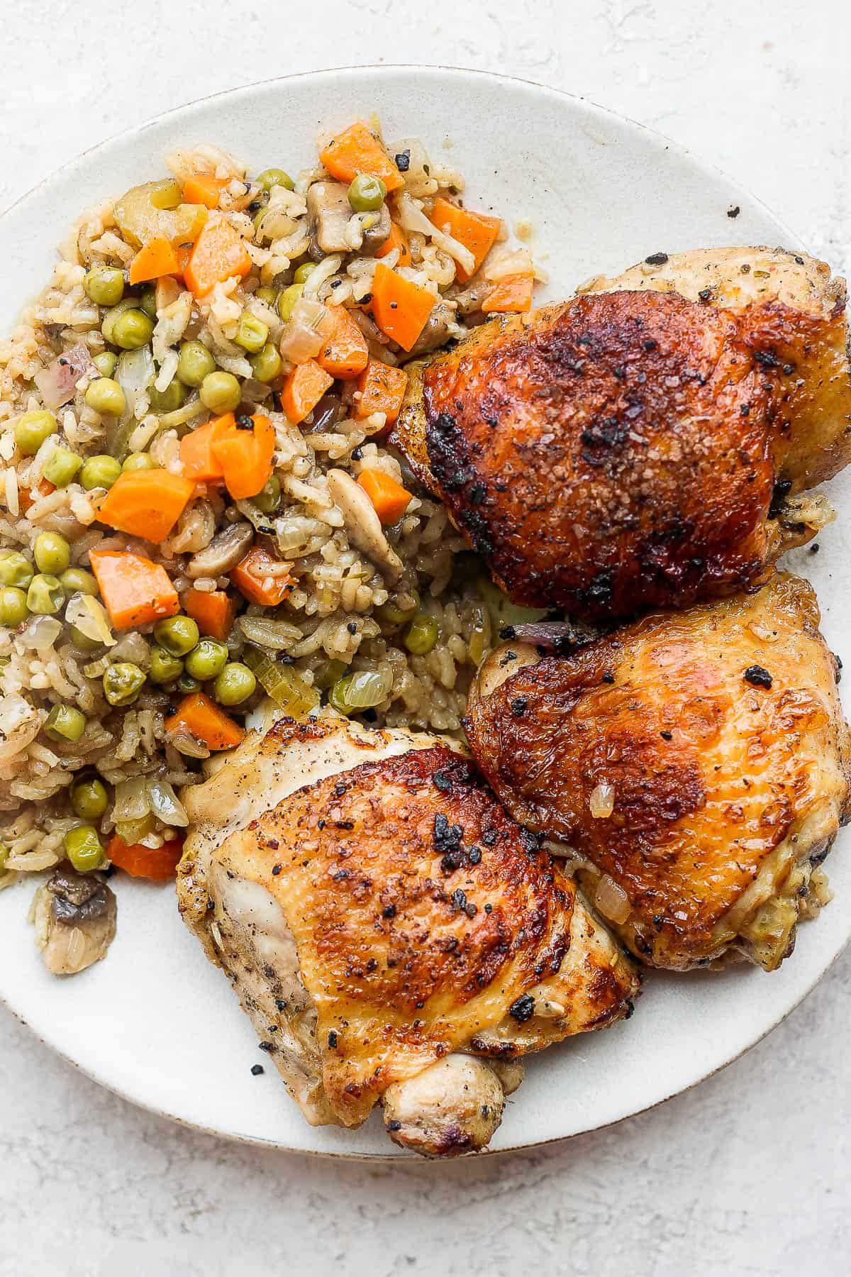 Three bone-in baked chicken thighs served with rice and veggies on a plate.