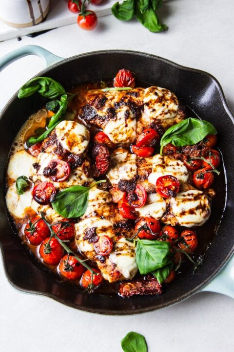 an overhead shot of caprese chicken with blistered grape tomatoes in a cast iron skillet garnished with fresh basil.