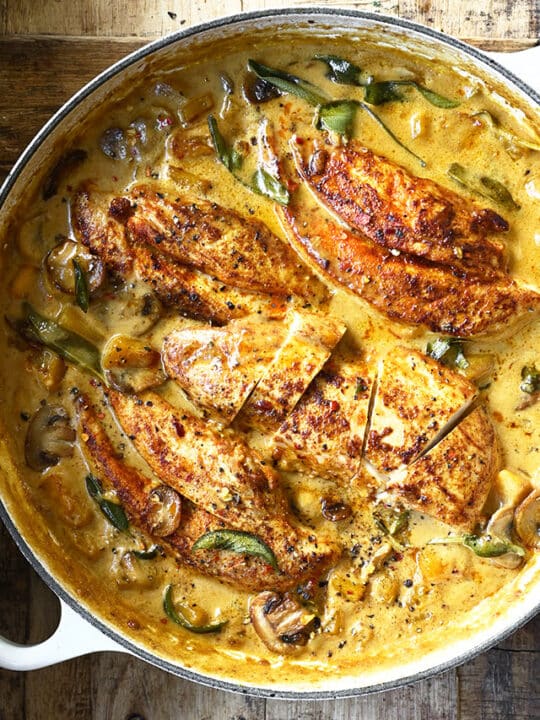 an overhead shot of seared chicken breasts nestled in a a creamy sauce garnished with sage leaves.
