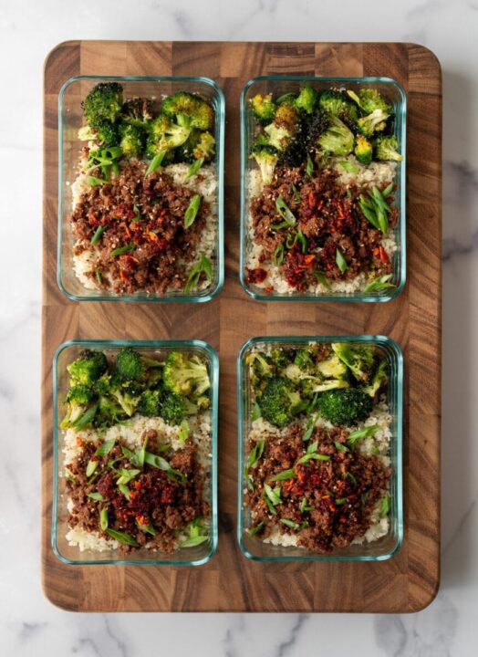 an overhead shot of four glass meal prep containers on a wooden cutting board filled with broccoli and ground bison.