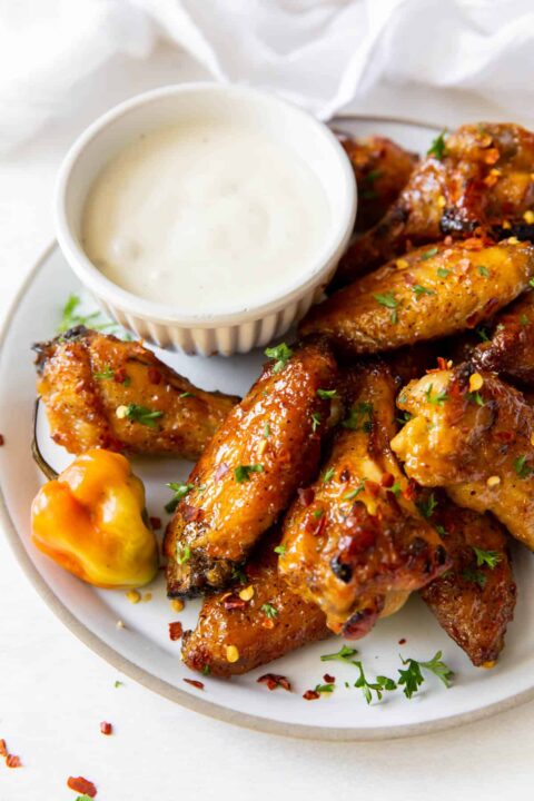 mango habanero chicken wings on a white plate beside a white dipping sauce and an habanero pepper sitting on the plate.