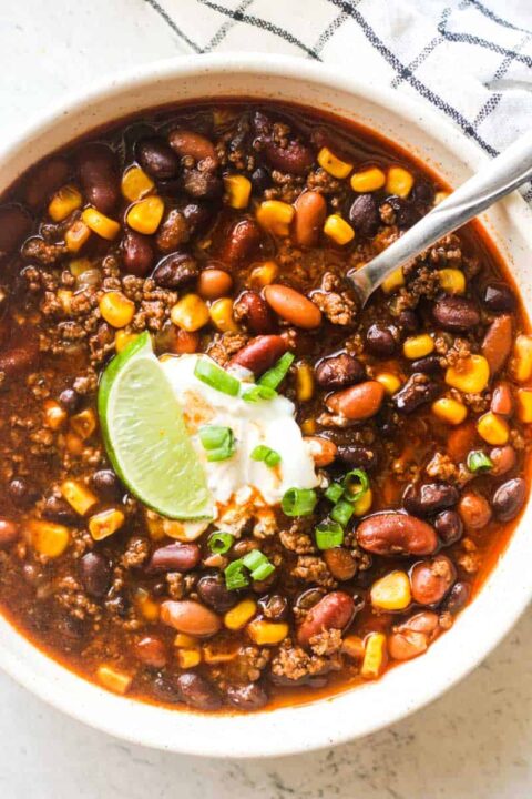 an overhead shot of bison chili in a white bowl topped with a dollop of sour cream and some sliced green onion and a wedge of lime.