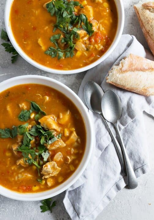 an overhead shot of two white bowls filled with pumpkin chicken soup and garnished with parsley and two silver spoons lying on a napkin beside the bowls.