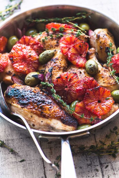 nicely browned chicken breast in a metal skillet with slices of blood oranges and olives nestled amongst the chicken.