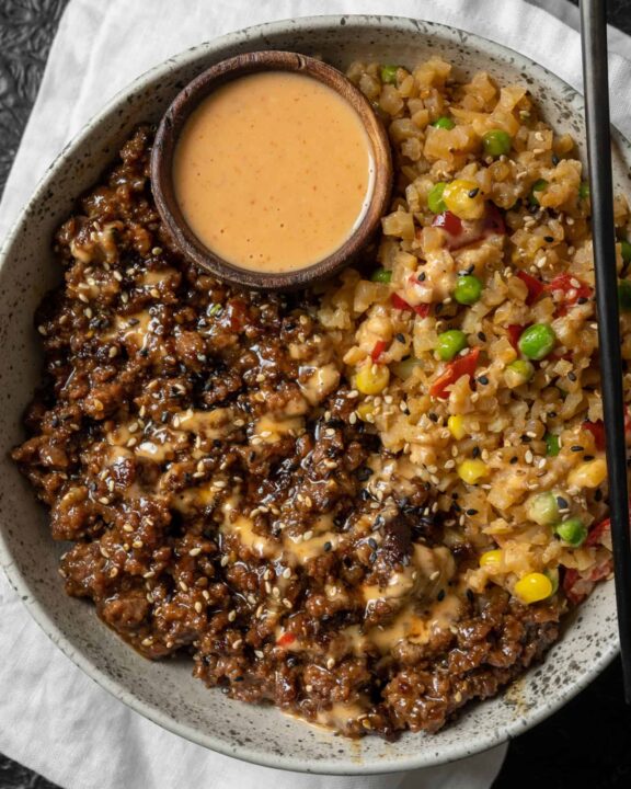 an overhead shot of teriyaki ground bison and rice in a large bowl with a small bowl of sauce resting on the rice.