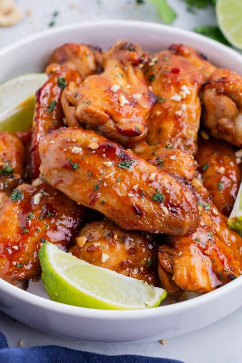 a close up shot of thai chicken wings in a white bowl with lime wedges tucked beside the wings.