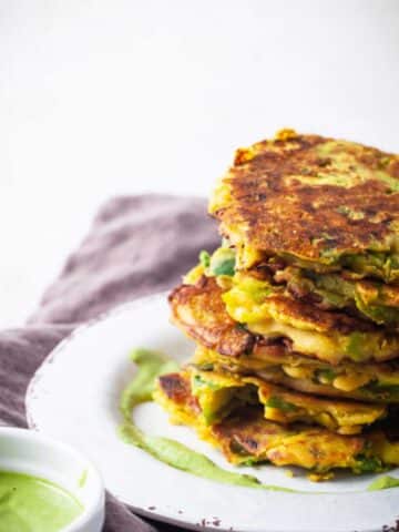 A stack of crispy leek veggie cakes served on a plate with a pesto sauce on the side.