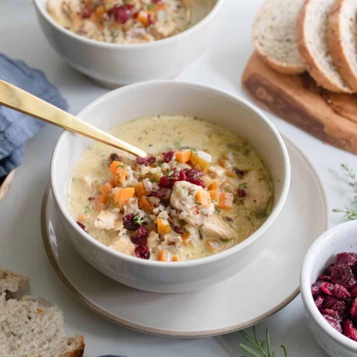 a bowl of wild rice cranberry soup in a white bowl with a gold spoon tucked in the side a loaf of sliced bread in the background.